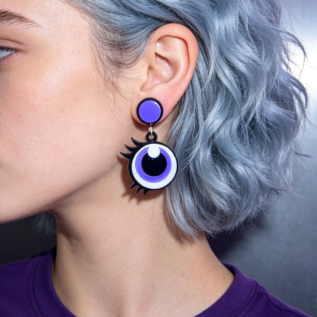 Purple cartoon eye dangle earrings on blue-haired model, black pupil with white highlight